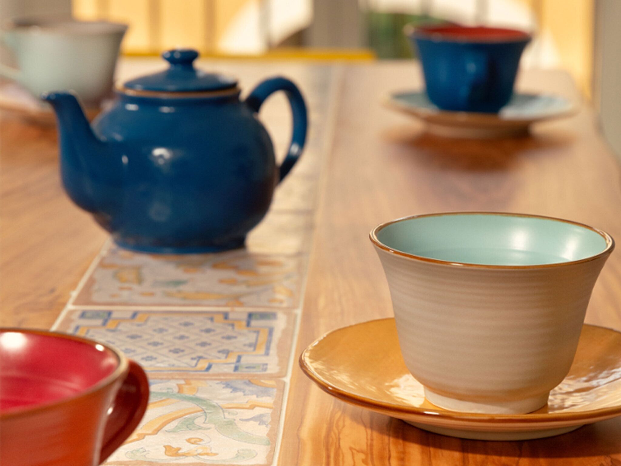 Una mesa con una tetera azul, una taza blanca sobre un platillo y una taza roja.