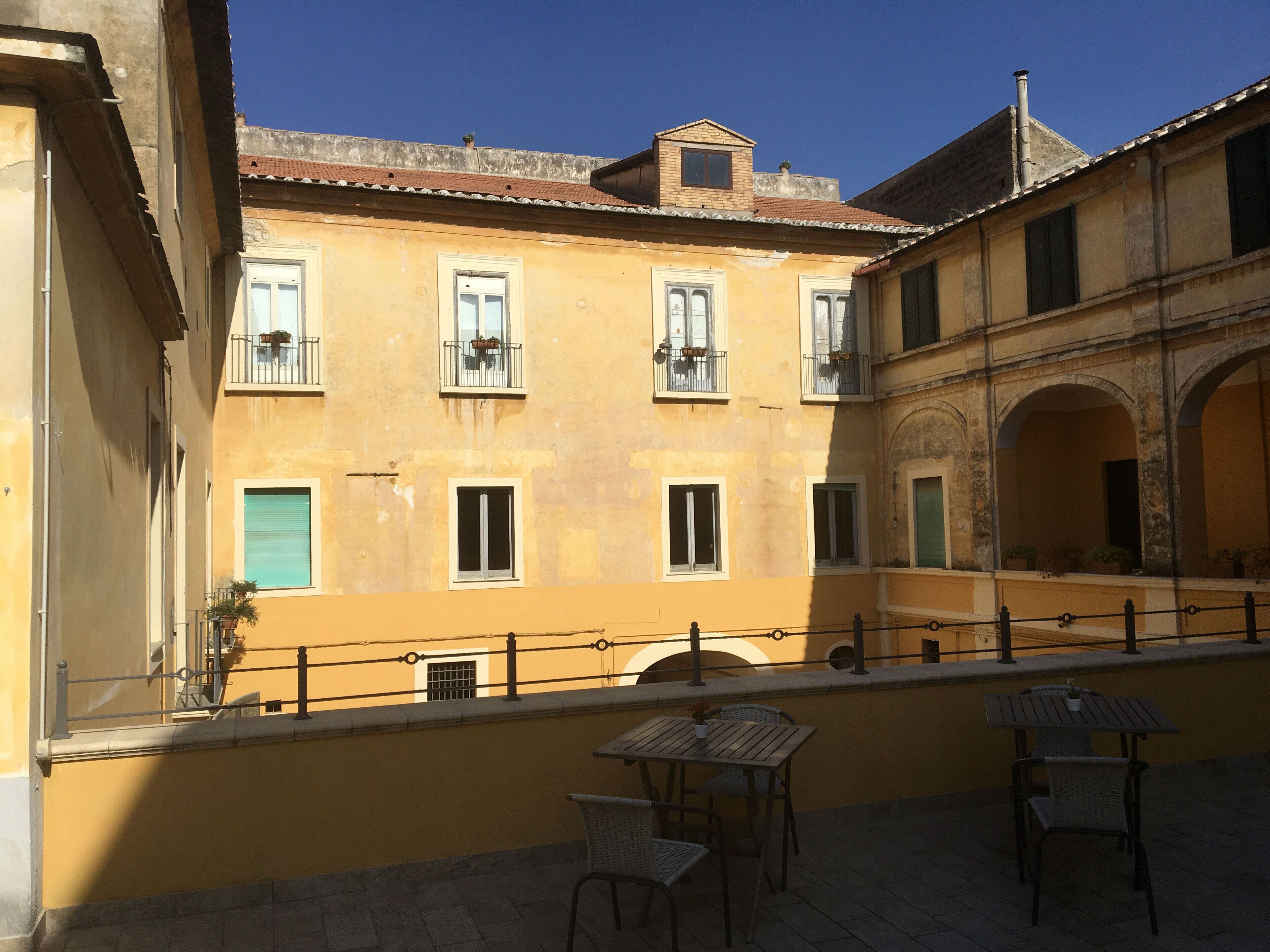 A courtyard with a yellow building, outdoor seating, and a railing.