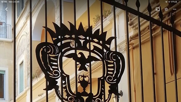 A wrought iron gate with a decorative emblem, set against a building facade with arches and a balcony. - Palazzo Benucci - Residenza di Charme (Santa Maria Capua Vetere)