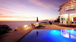 Zona piscina con vista sull'oceano al tramonto, arredi esterni e un edificio con balcone.