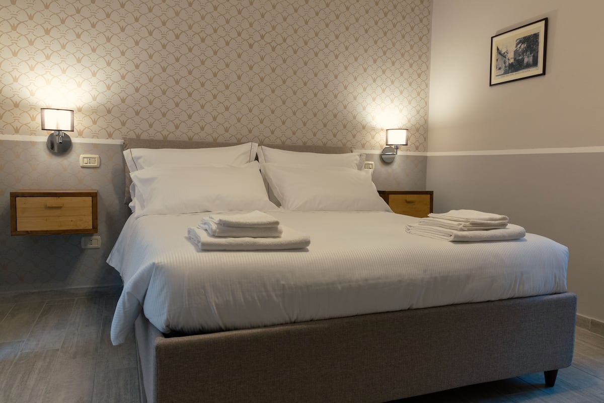 A neatly made bed with white linens and towels, two bedside tables with lamps, a wooden nightstand, and a patterned wallpaper.