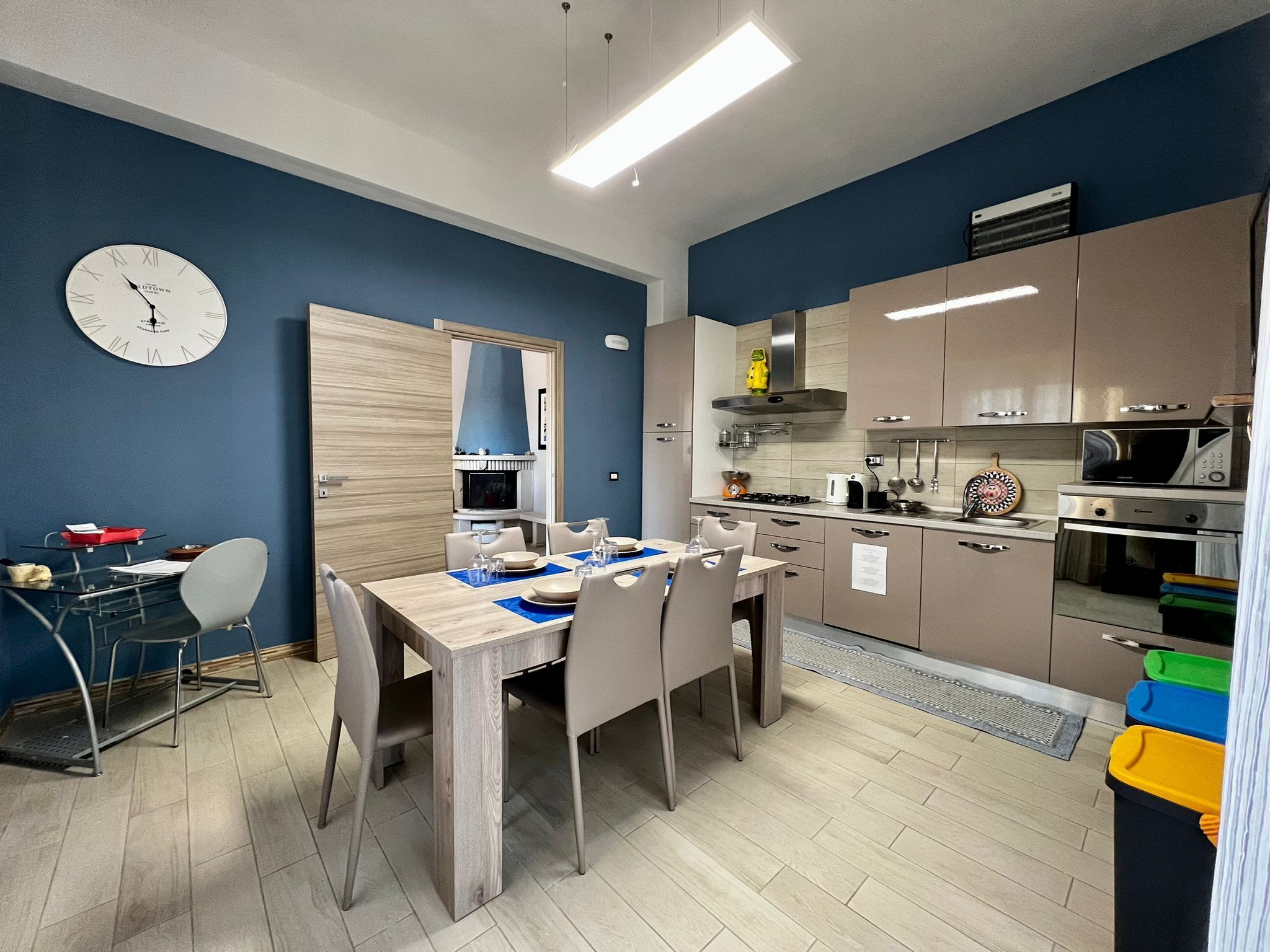 A modern kitchen with stainless steel appliances, a dining area with a wooden table and chairs, and a blue accent wall with a clock.