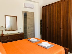 A hotel room with a wooden wardrobe, a bed with an orange bedspread, a mirror, a lamp, and a wall-mounted air conditioner. - Eco-friendly Holiday Apartment Marina di Ragusa apt "il Gufo" (Ragusa)