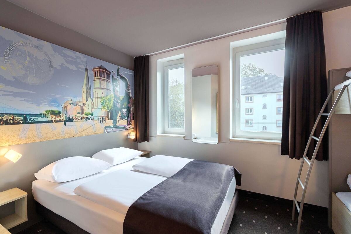 A hotel room with a bed, a nightstand, a window with curtains, a wall-mounted mirror, and a painting of a cityscape.