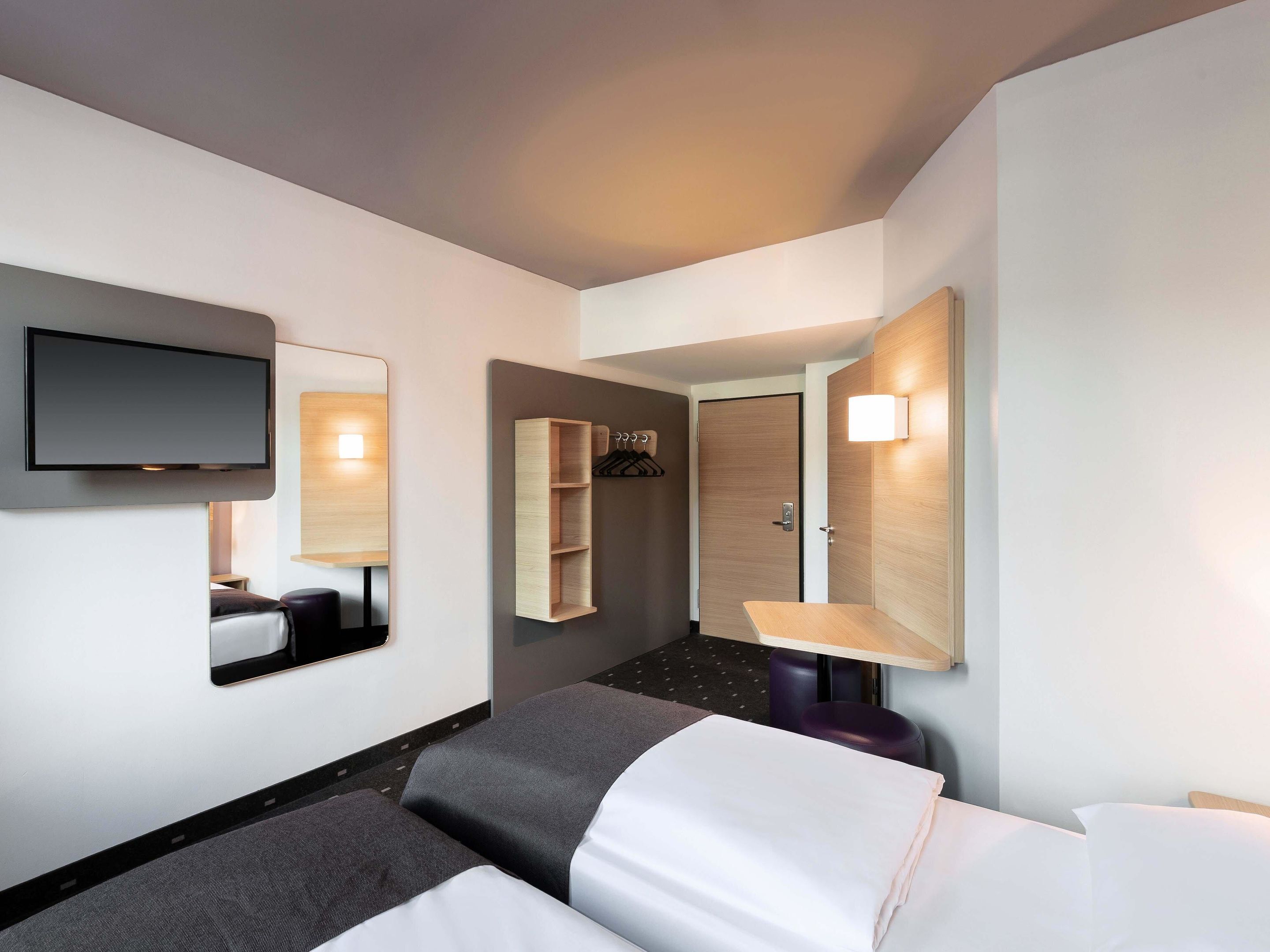 Une chambre d’hôtel comprenant un lit, un téléviseur à écran plat fixé au mur, un miroir, une étagère en bois, une porte et une petite table avec une lampe.
