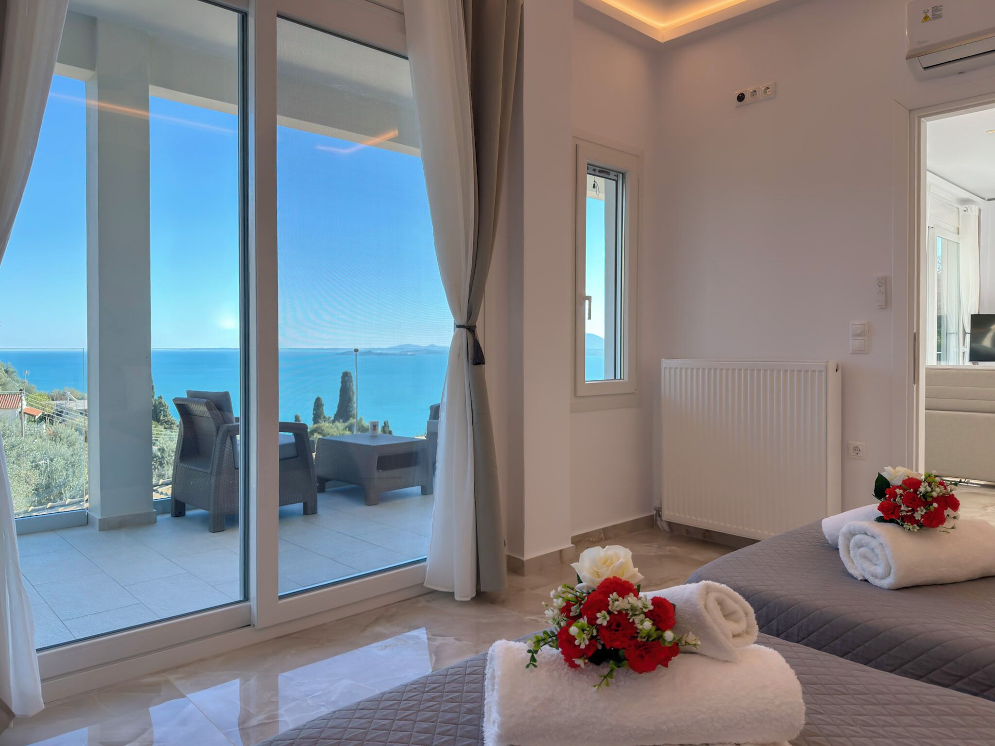 A hotel room with a large sliding glass door leading to a balcony with a view of the sea. There are two beds with towels and a bouquet of flowers.