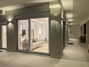 Um interior moderno com uma porta de vidro deslizante que dá para uma sala de estar, uma cozinha com armários brancos e um corredor com sofá e poltronas.
