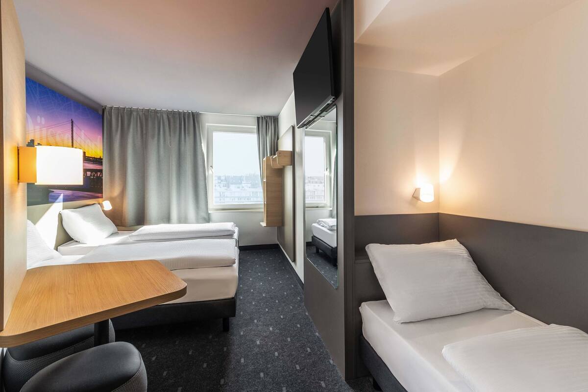 Habitación de hotel con cama, escritorio, silla, ventana con cortinas, televisor montado en la pared y vista panorámica de la ciudad.