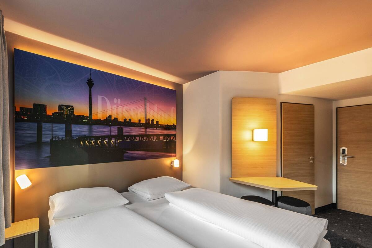 Habitación de hotel con cama, escritorio, lámpara de pared y un cuadro de un paisaje urbano.