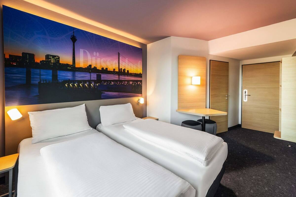Habitación de hotel con dos camas, un mural de paisaje urbano, una mesita, una mesita de noche y una puerta.