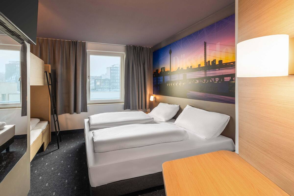 Habitación de hotel con dos camas, un escritorio, una silla, un ventanal con cortinas y un cuadro de paisaje urbano en la pared.