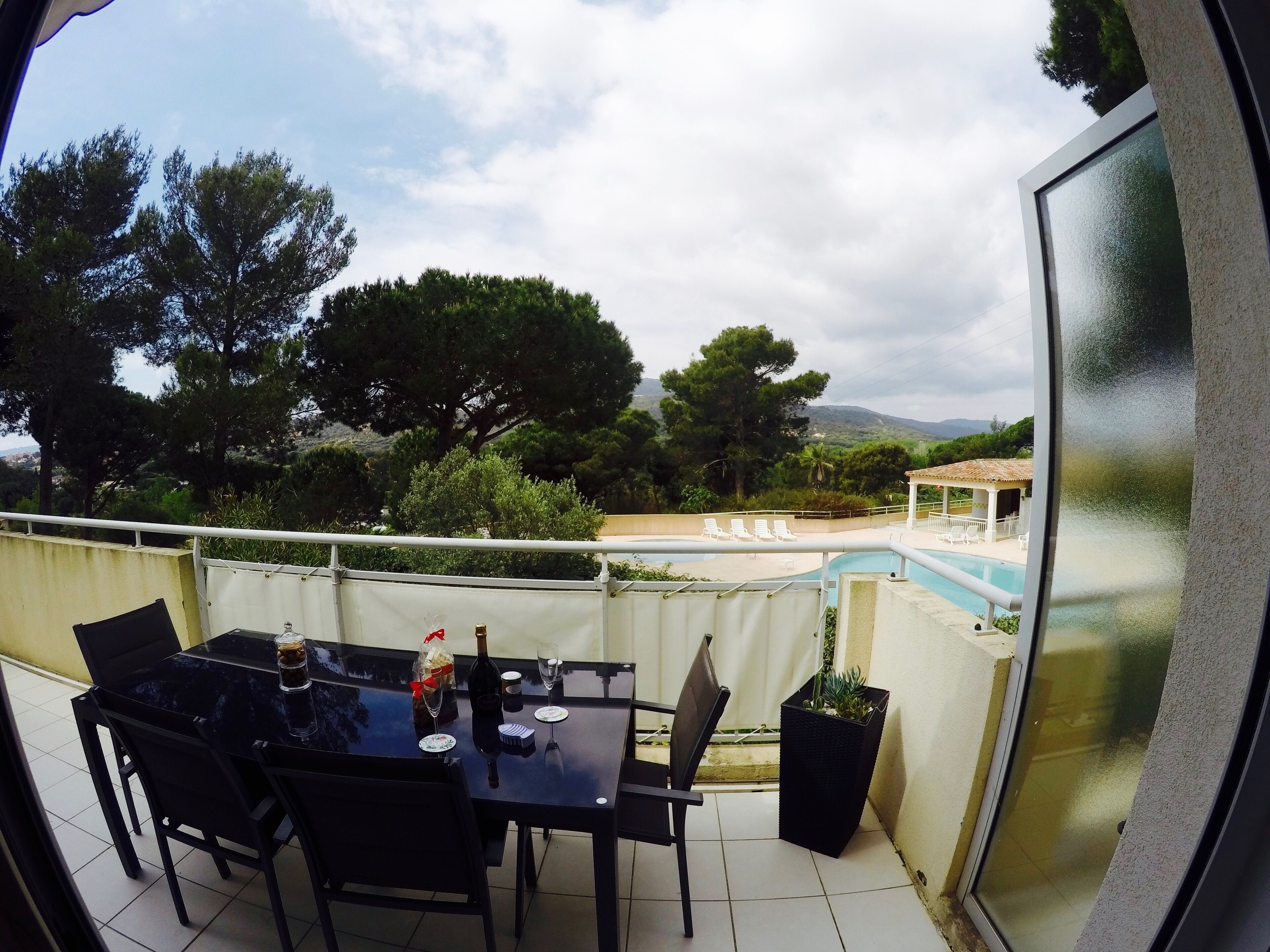 A balcony with a table set for two, overlooking a swimming pool and a garden.