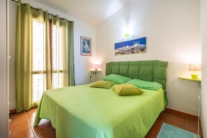A bedroom with a bed, curtains, a wall-mounted lamp, a painting, and a tiled floor.