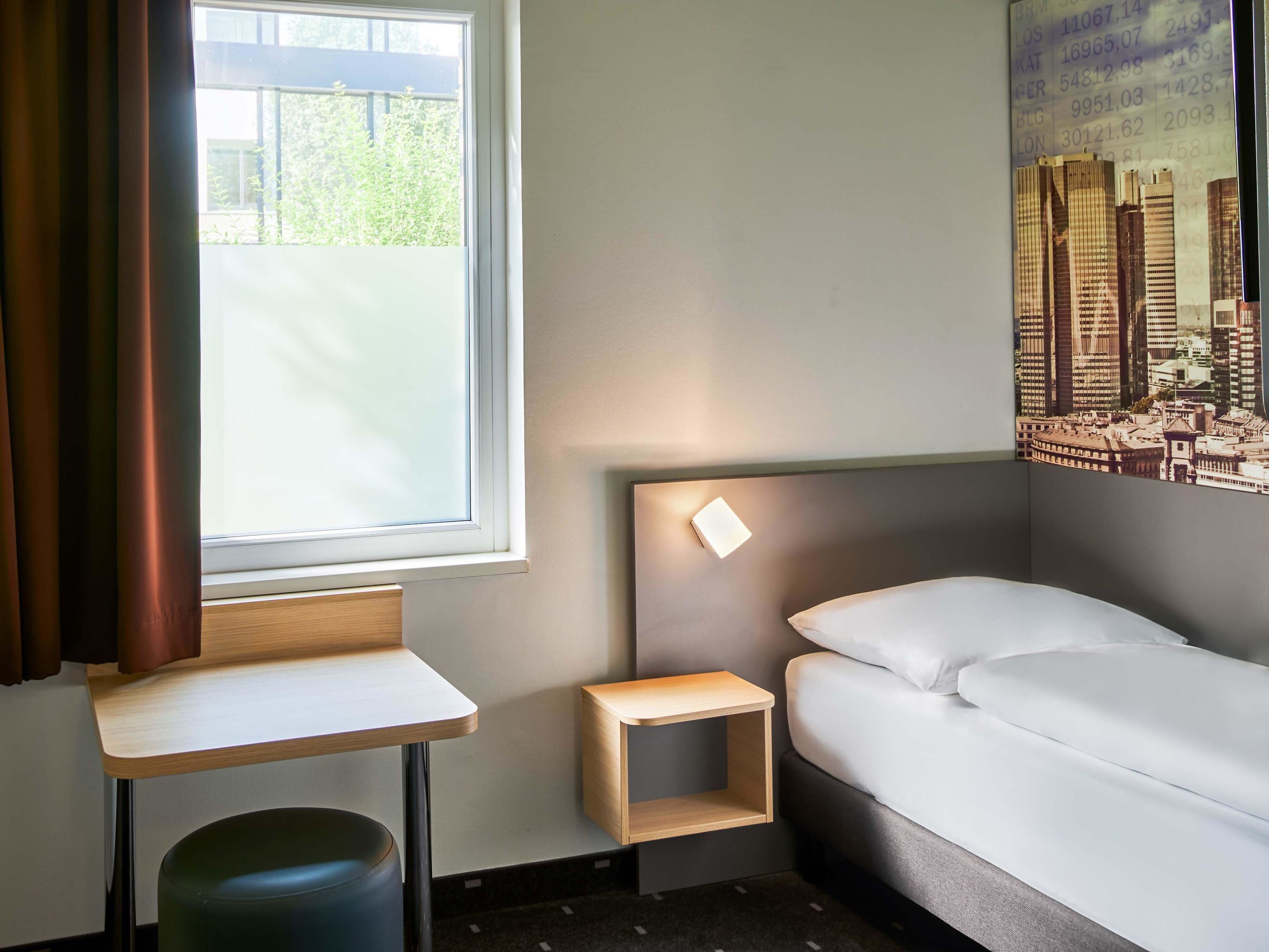 Una habitación de hotel con cama, una mesita pequeña, una mesilla de noche, una ventana con cortinas y un cartel con vistas a la ciudad en la pared.