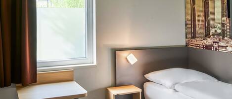 Una habitación de hotel con cama, una mesita pequeña, una mesilla de noche, una ventana con cortinas y un cartel con vistas a la ciudad en la pared.