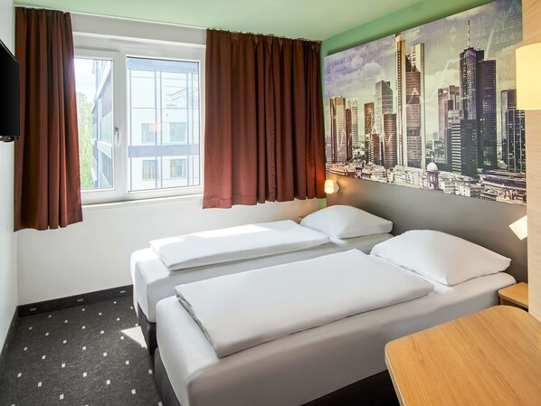 Una habitación de hotel con dos camas, una ventana con cortinas marrones, un mural de paisaje urbano y una pequeña mesa de madera.