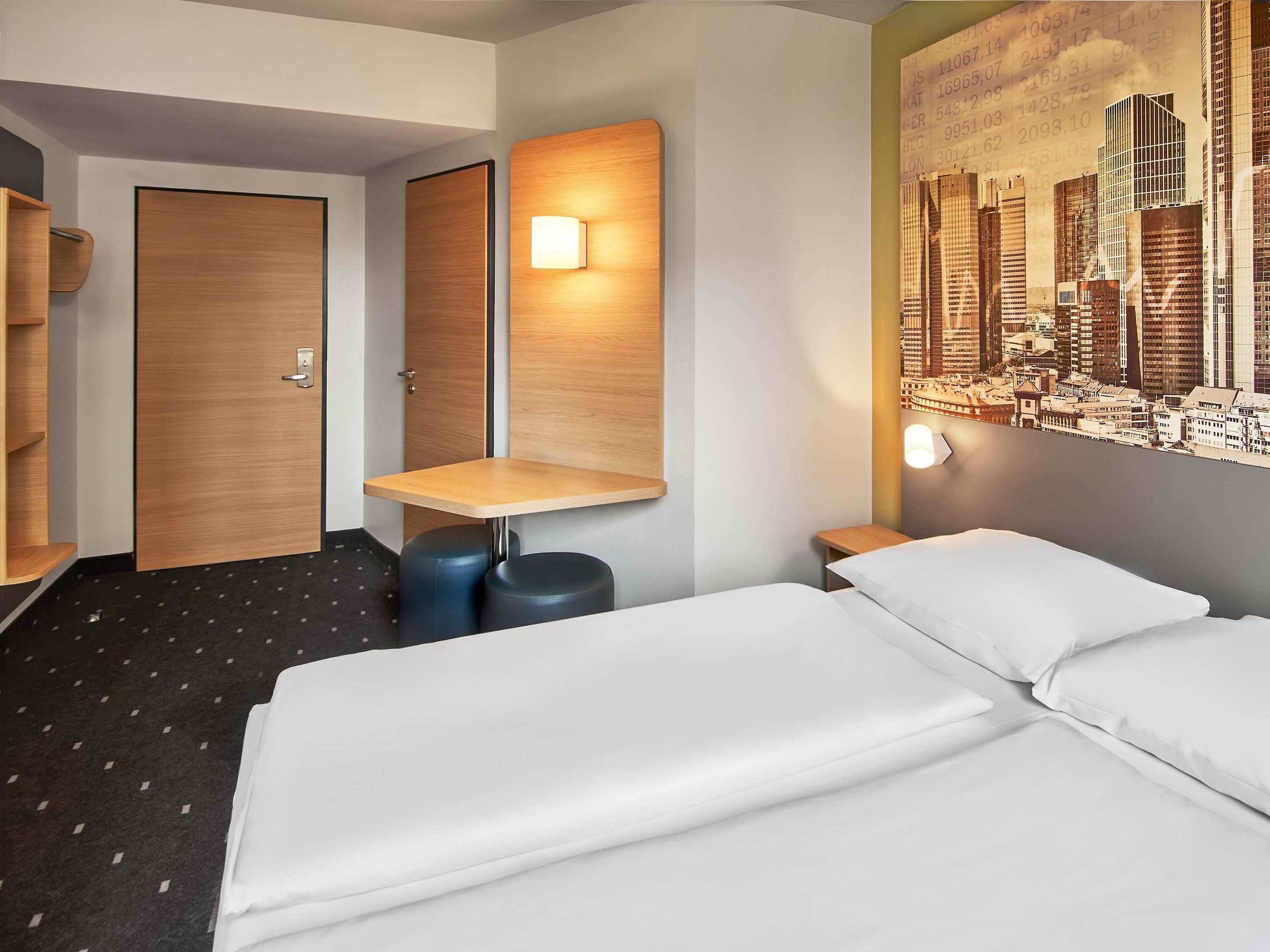 Una habitación de hotel con cama, escritorio, dos taburetes, una puerta y una lámpara de pared.