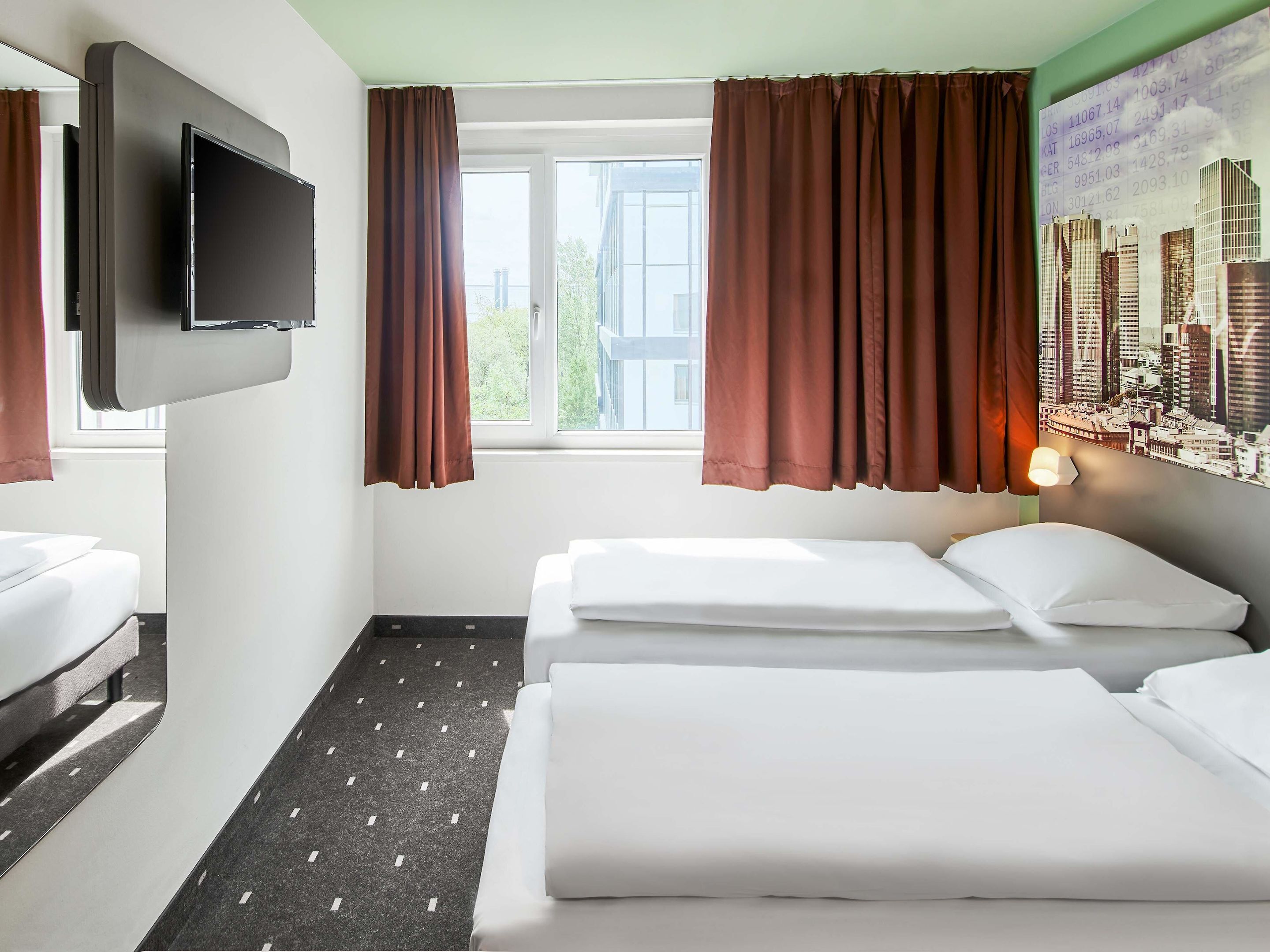 Habitación de hotel con dos camas, un televisor de pantalla plana montado en la pared y una ventana con cortinas marrones.