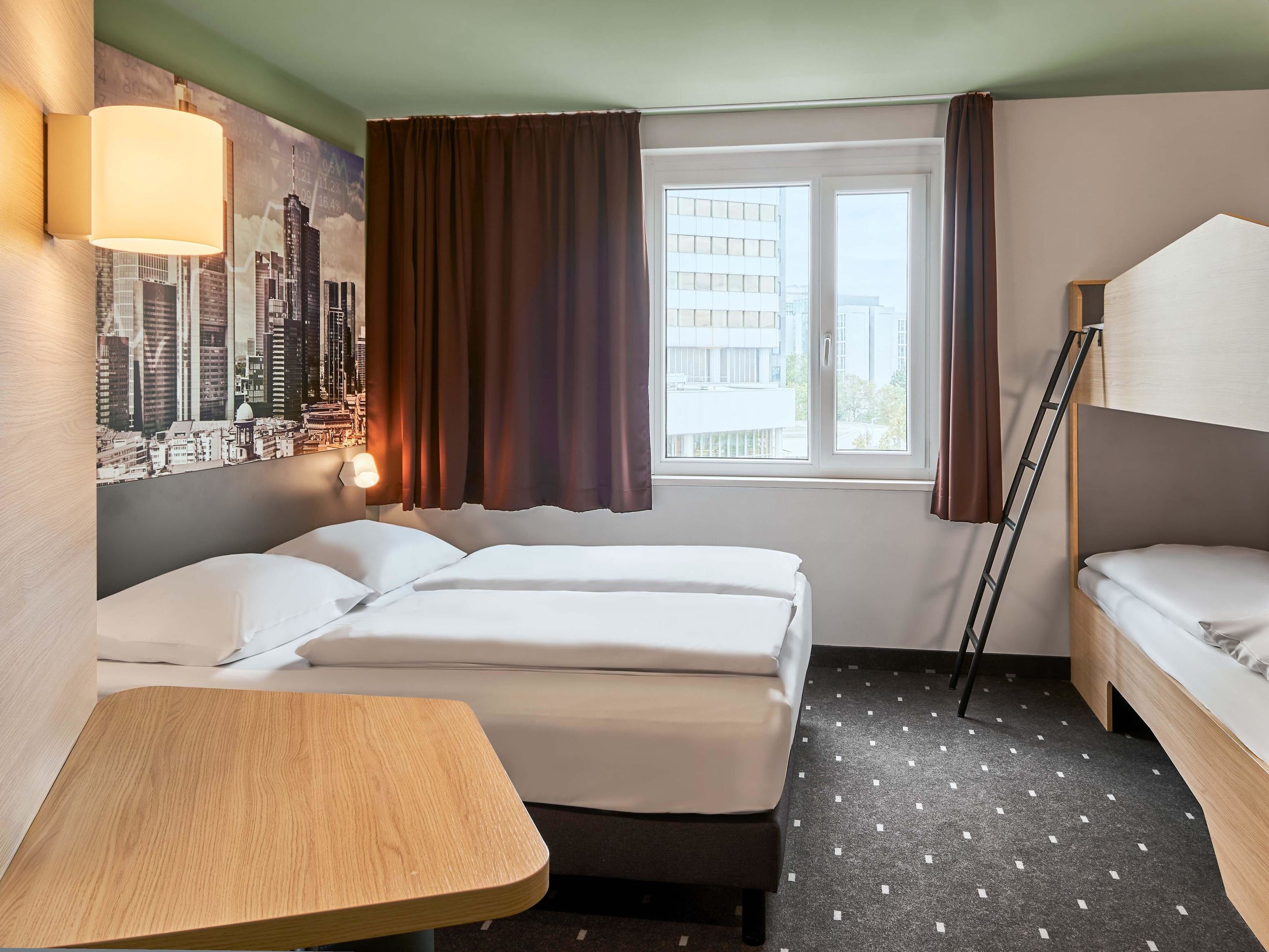 A hotel room with two beds, a wooden table, a cityscape mural, a window with curtains, and a view of buildings.