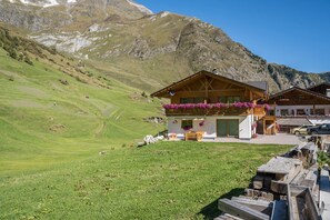 Un rifugio di montagna con facciata in legno, un balcone adornato da fiori pendenti e vista sulle colline circostanti.