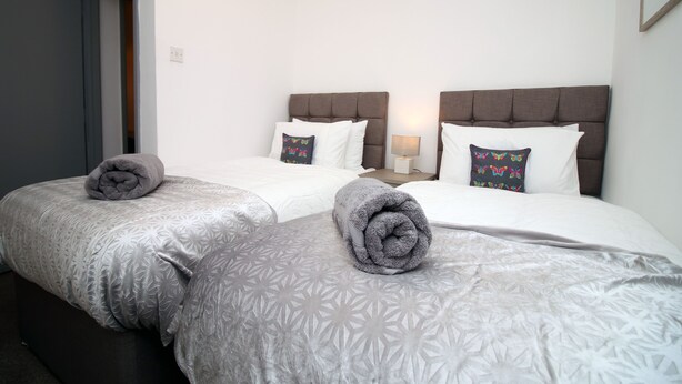 A hotel room with two beds, each with a grey patterned bedspread and pillows, a nightstand with a lamp, and a door leading to another room. - BOOM Swansea City (Swansea)
