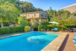 Une piscine à l’eau claire, une maison en pierre dotée d’un balcon et une végétation luxuriante tout autour.