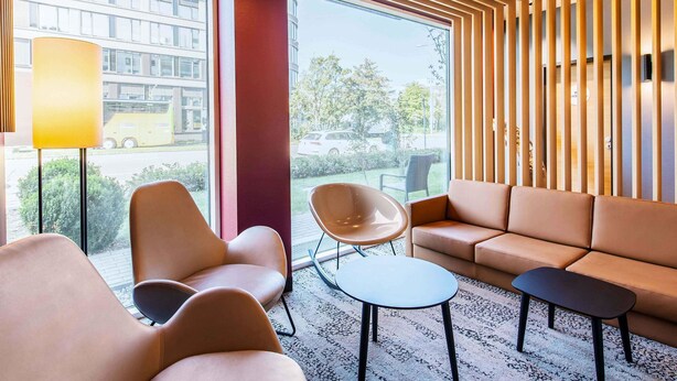 A modern lounge area with leather sofas, a round table, and a lamp. Large windows provide a view of the street and buildings outside.