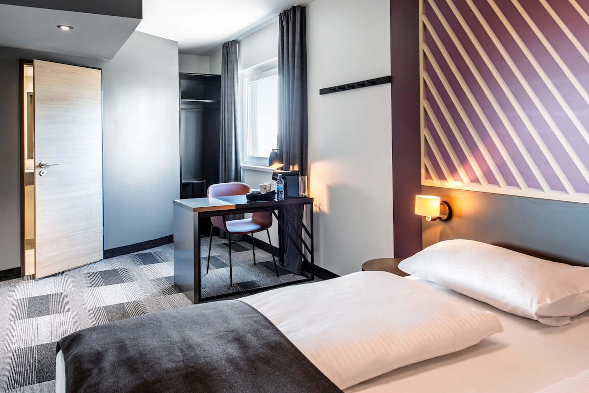 A hotel room with a bed, a desk with a chair, a lamp, and a window with curtains.