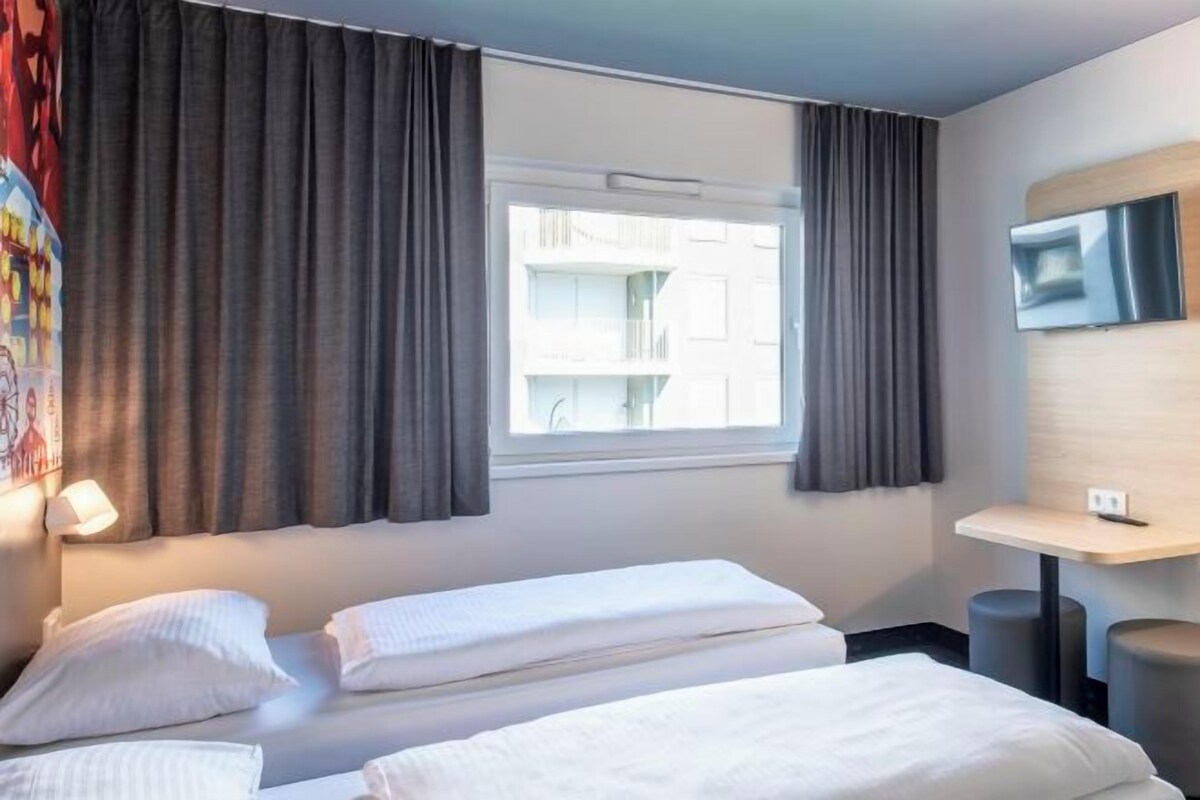 A hotel room with two beds, a TV mounted on the wall, a window with curtains, and a small table with a lamp.