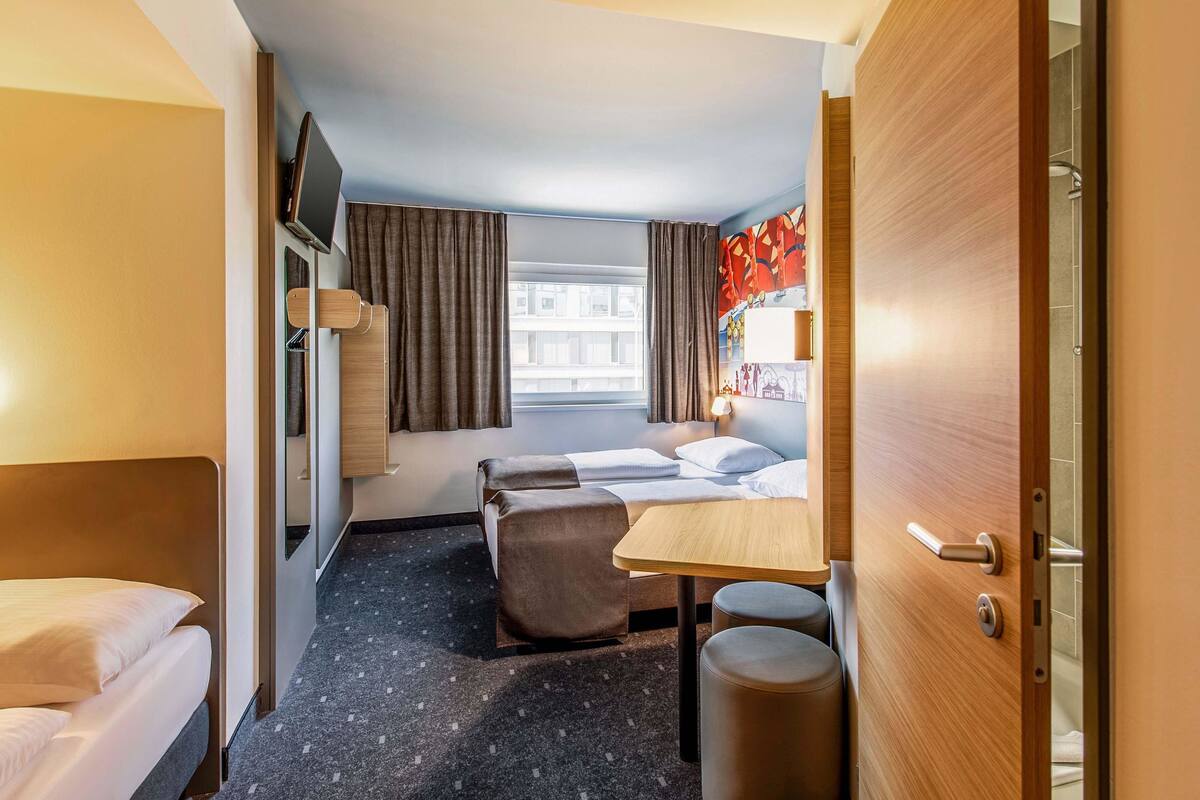 A hotel room with a bed, a desk, a chair, a small table, a TV mounted on the wall, and a window with curtains.
