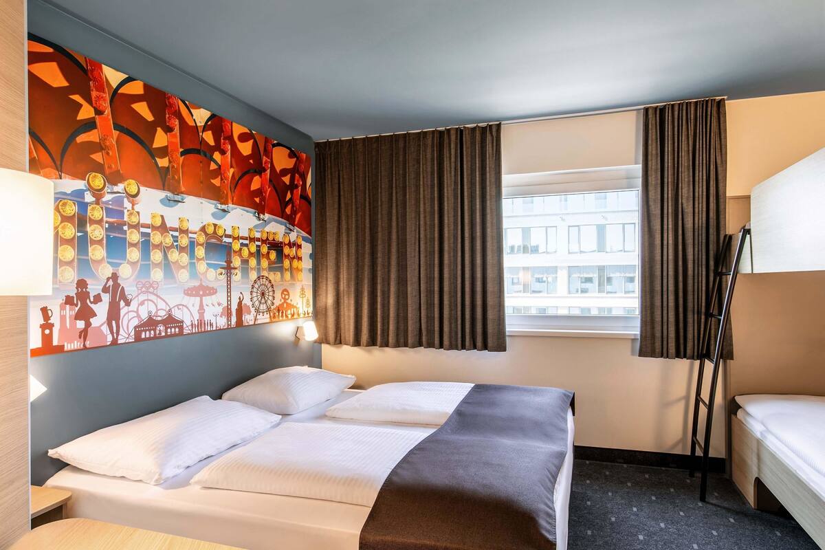 A hotel room with a bed, a desk, a chair, a window with curtains, and a wall with a colorful mural.