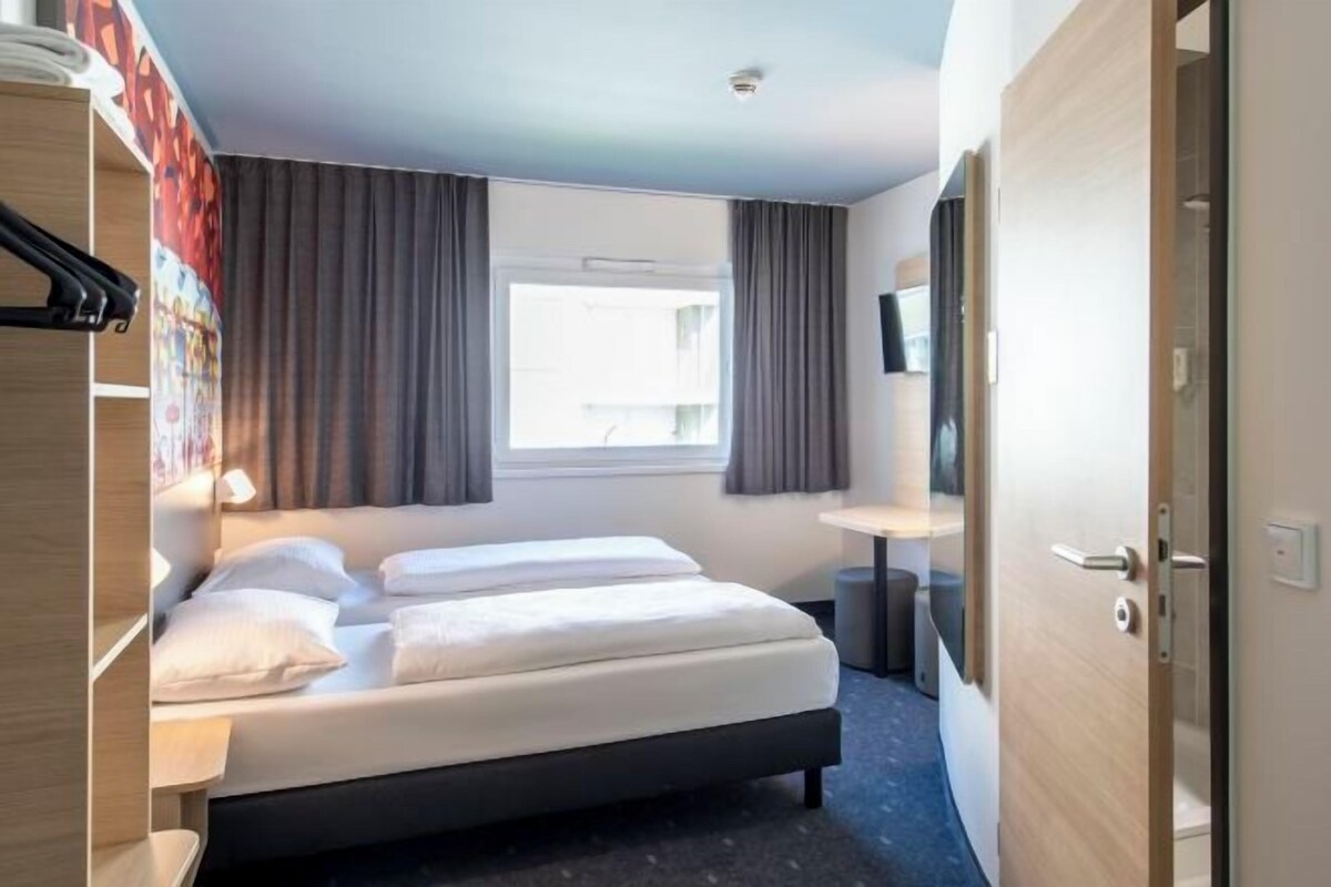 A hotel room with a bed, a desk, a chair, a wardrobe, and a window with curtains.