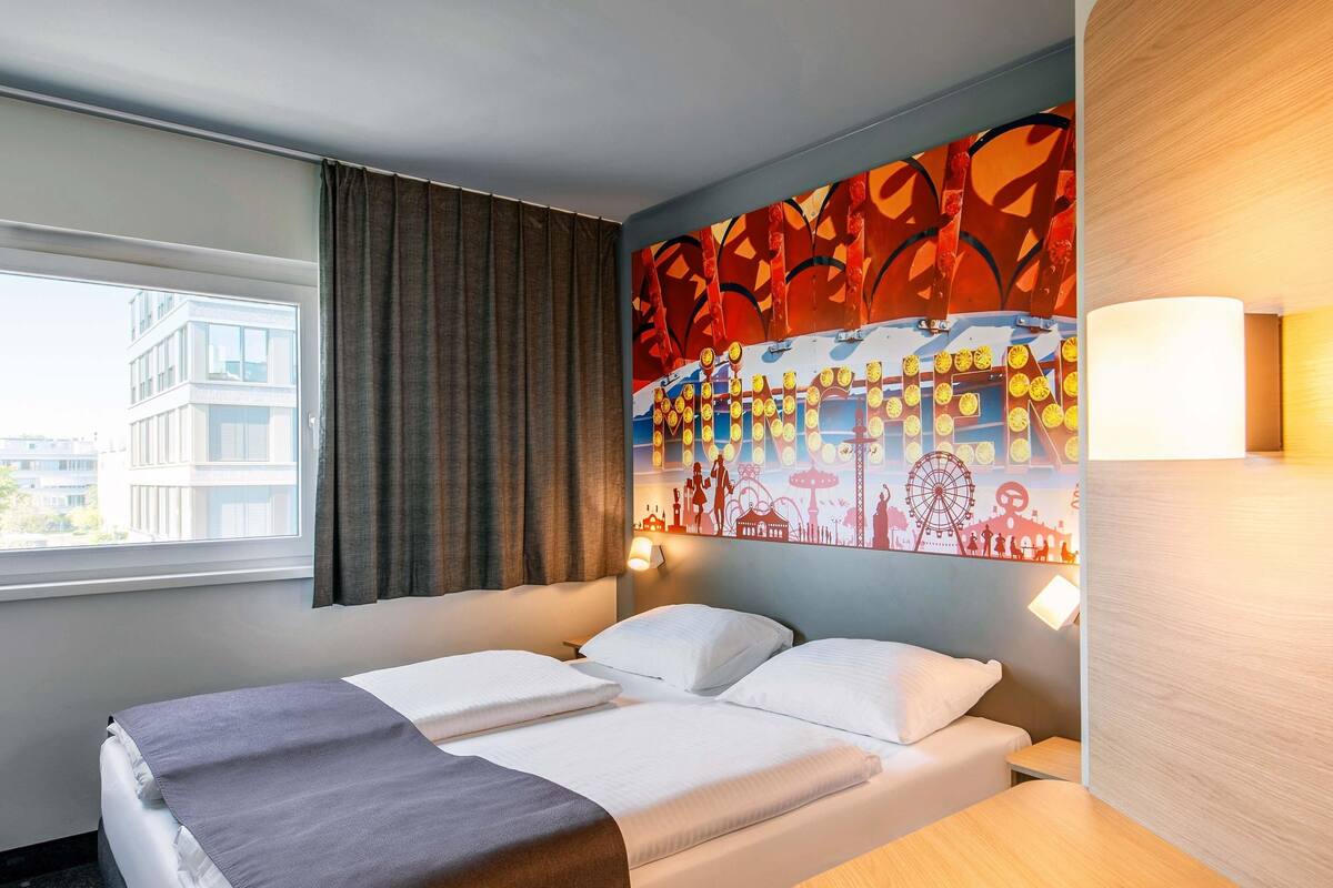 A hotel room with a bed, a desk, a window with curtains, and a wall mural.