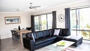 A modern living room with a black leather sofa, a glass coffee table, a dining table with chairs, and large windows with curtains.