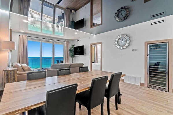 Una habitación de hotel moderna con una gran mesa de comedor de madera, asientos cómodos, un balcón con vista al mar y un televisor montado en la pared.