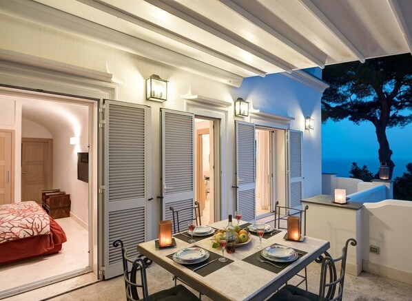 A dining area with a table set for four, a view of the sea, and a bedroom with a red bedspread.