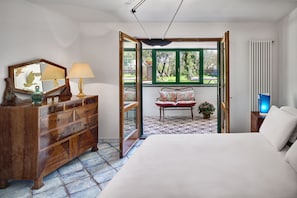 A bedroom with a bed, a dresser, a mirror, a lamp, and a view of a garden through an open door. - Stony Villa Volcano View (Massa Lubrense)