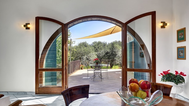 A dining area with a glass table, chairs, and a view through a large arched doorway to an outdoor patio with a table and chairs. - Stony Villa Volcano View (Massa Lubrense)