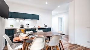 Une cuisine moderne avec une table à manger en bois et des chaises blanches, un téléviseur à écran plat fixé au-dessus du comptoir, et une vue sur la pièce adjacente.