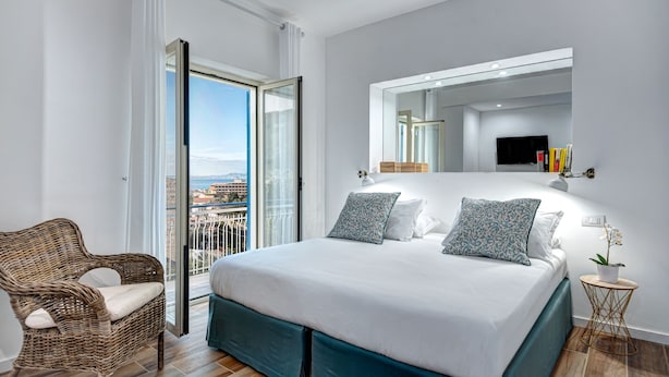 Une chambre moderne avec un grand lit, un fauteuil en osier, un balcon avec vue sur la ville et un téléviseur fixé au mur.