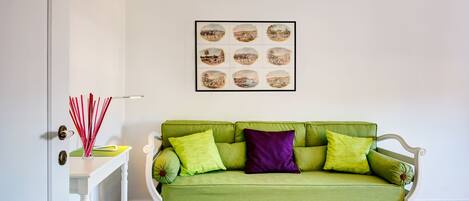 A green sofa with multiple cushions, a white side table with magazines, and a framed picture above the sofa.