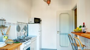 A compact kitchen with a gas stove, microwave, and a refrigerator. There is a wooden table with a loaf of bread and a green cutting board. A door with a frosted glass panel is visible, and a small table with a bottle and a glass is on the right.