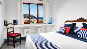 A bedroom with a large bed, a wooden chair, a window with curtains, and a view of a coastal town.