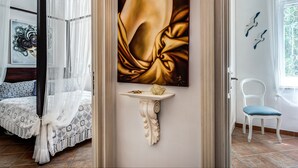 Une chambre à coucher avec un lit à baldaquin, un tableau au mur, une petite étagère ornée d’un objet décoratif et une chaise près de la fenêtre.