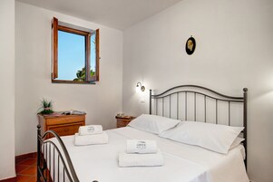 Cama bem arrumada com lençóis brancos e duas toalhas, penteadeira de madeira com uma planta, mesa de cabeceira com abajur e janela com uma persiana aberta.