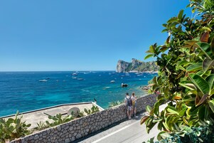 Uma estrada costeira com um muro de pedra e vegetação, que leva a uma vista do mar com barcos e um penhasco rochoso.