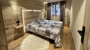 A bedroom with a wooden headboard, a patterned bedspread, a fluffy rug, and a window with curtains. - BOREAL - Lovely apartment in the village center and close to the slopes - Wi-Fi - Garage (La Clusaz)