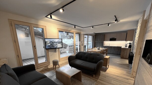A modern living room with a sofa, ottoman, and a fluffy rug. There is a television mounted on the wall and a kitchen area in the background. - BOREAL - Lovely apartment in the village center and close to the slopes - Wi-Fi - Garage (La Clusaz)