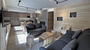 Un soggiorno moderno con un divano grigio, un tavolino da caffè e una TV. Sullo sfondo, una cucina con mobili in legno e una parete decorata con pietra.