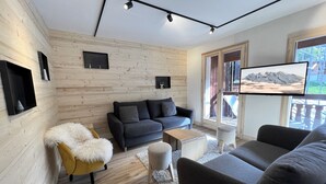 Un soggiorno moderno con un divano grigio scuro, una poltrona bianca e soffice e un tavolino in legno. C'è una TV a schermo piatto montata a parete e una finestra con vista sulle montagne.